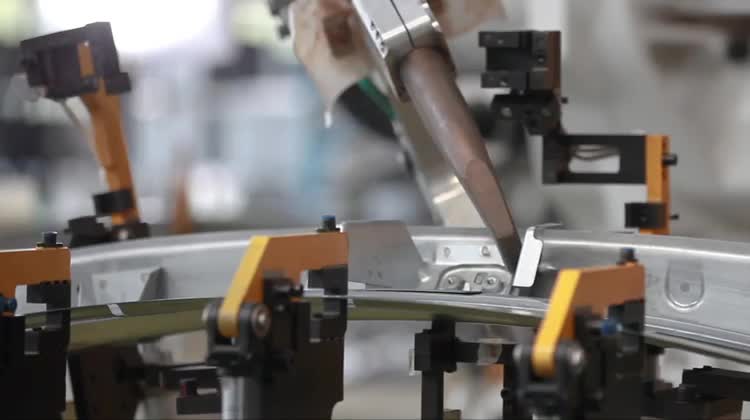 Close-up video frame of automated welding or joining activity on a metal component fixture.